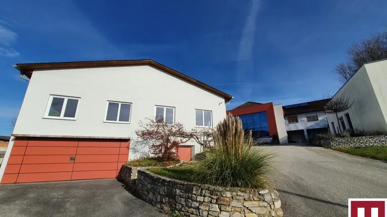 Gebäudeansicht mit Zufahrt und Garagentor unter blauem Himmel, umgeben von leichter Vegetation.