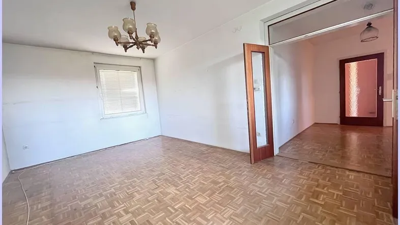 Helles, leeres Wohnzimmer mit Parkettboden und einer Tür, die zu einem weiteren Raum führt.