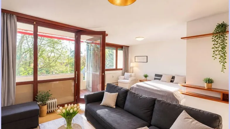 Helles Wohnzimmer mit Sofa, Bett und Zugang zum Balkon, umgeben von viel Grün.