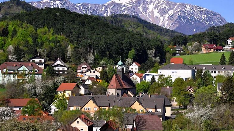 Malerisches Dorf mit traditionellen Häusern, umgeben von grünen Hügeln und schneebedeckten Bergen.
