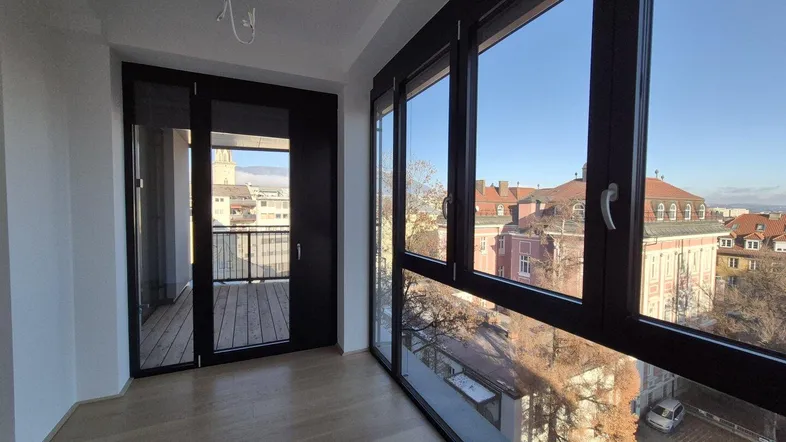 Heller Raum mit großen Fenstern, Holzboden und Zugang zu einem Balkon mit Stadtblick.