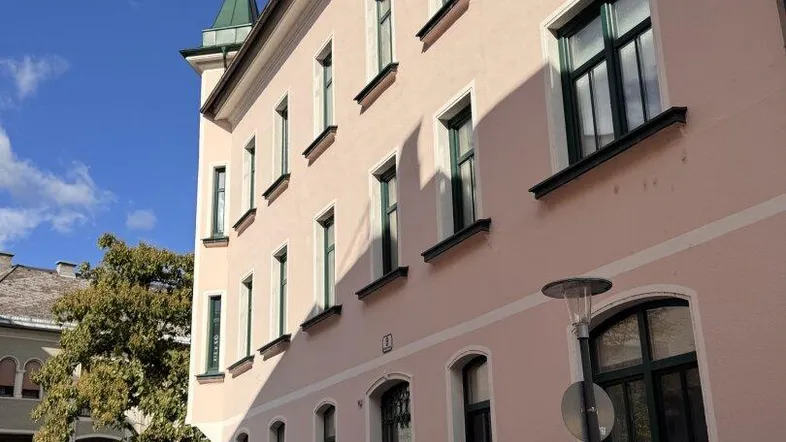 Historisches Gebäude mit hellrosa Fassade, grünen Fensterrahmen und einem markanten Eckturm.