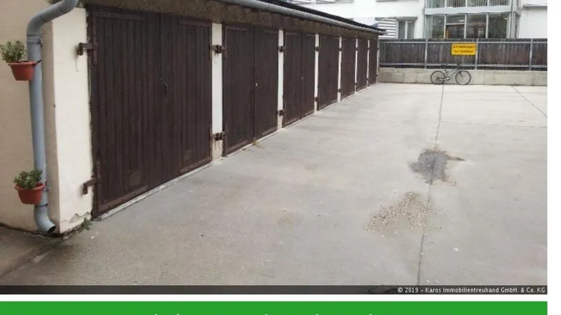 Reihe von Garagenboxen mit braunen Holztoren und einem Mehrfamilienhaus im Hintergrund auf einem gepflasterten Vorplatz.