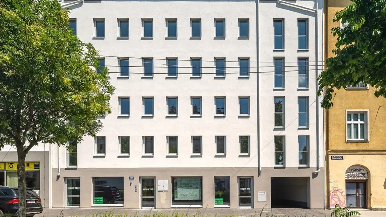 Modernes Mehrfamilienhaus mit weißer Fassade, vielen Fenstern und begrüntem Vorgarten in städtischer Umgebung.