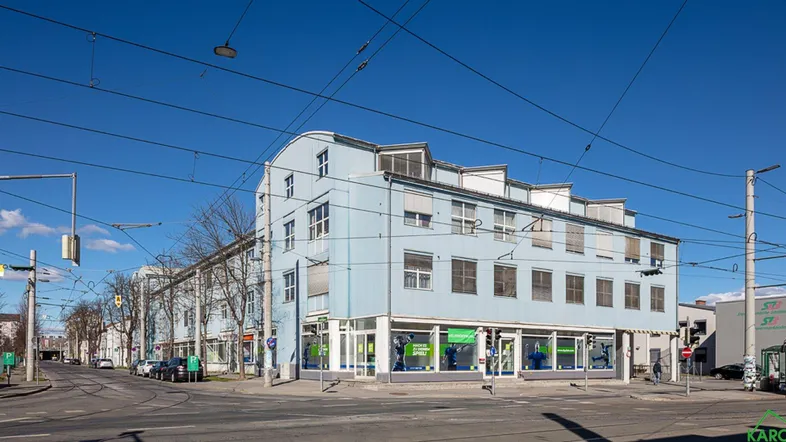 Gebäudeansicht von der Straße mit blauer Fassade und großen Fenstern im Erdgeschoss, umgeben von Oberleitungen.