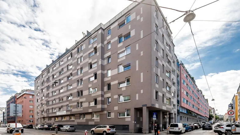 Großes Wohngebäude an einer Straßenecke mit vielen Fenstern und urbaner Umgebung.
