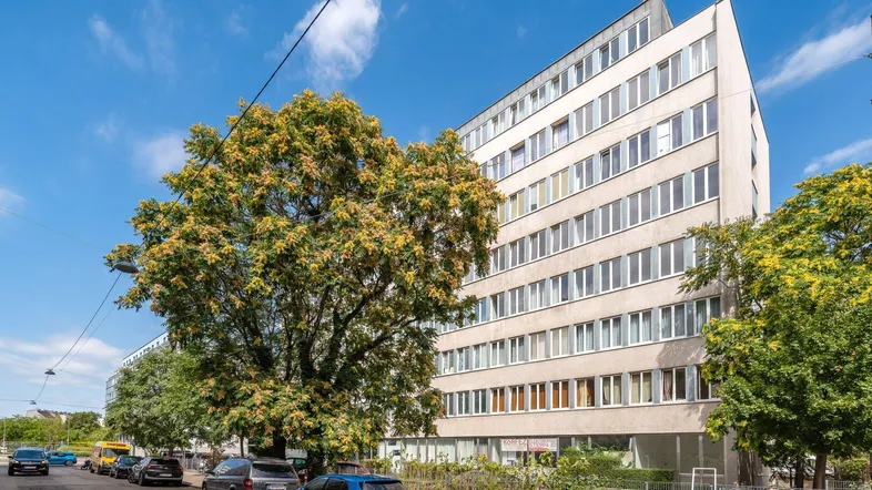 Mehrstöckiges Wohngebäude mit vielen Fenstern und Bäumen an einer städtischen Straße.