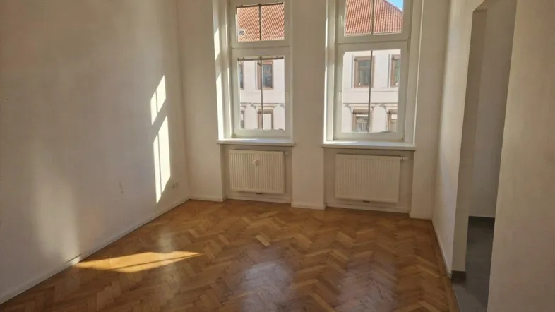 Helles Zimmer mit Fischgrätparkettboden und zwei großen Fenstern mit Blick nach draußen.