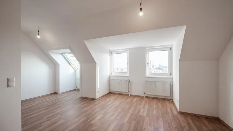 Helles Zimmer im Dachgeschoss mit schrägen Wänden, Laminatboden und Blick nach draußen.