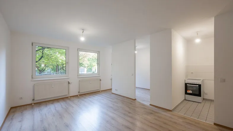 Heller Wohnraum mit Laminatboden, großen Fenstern und Blick ins Grüne, inklusive einer kleinen Küchenzeile.