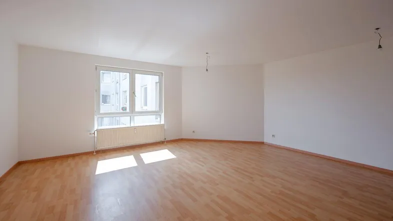 Helles, leeres Wohnzimmer mit Laminatboden und großem Fenster, das viel Tageslicht hereinlässt.