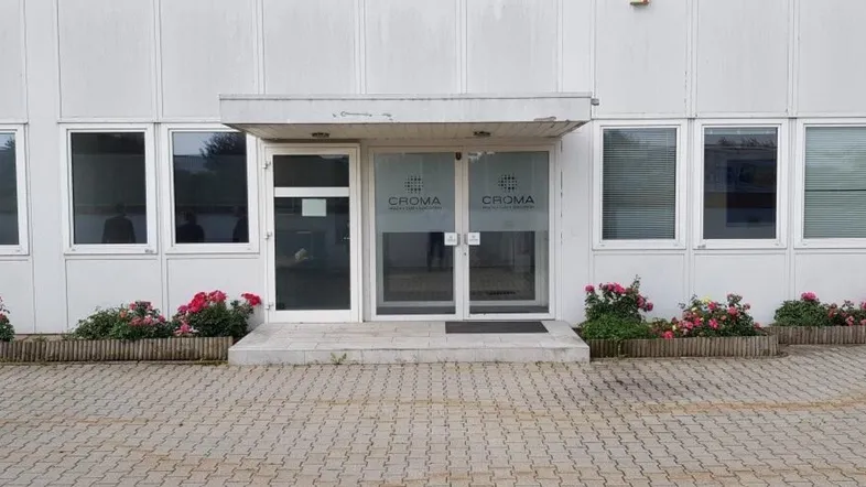 Eingangsbereich eines Geschäftsgebäudes mit weißer Fassade, Glastüren und kleinen Blumenbeeten.
