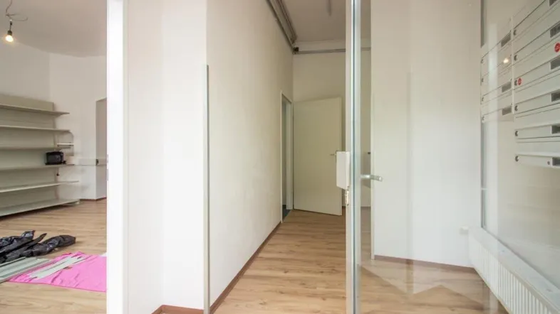 Heller Eingangsbereich mit Laminatboden, Glaswand und Blick in einen angrenzenden Raum mit Regalen.
