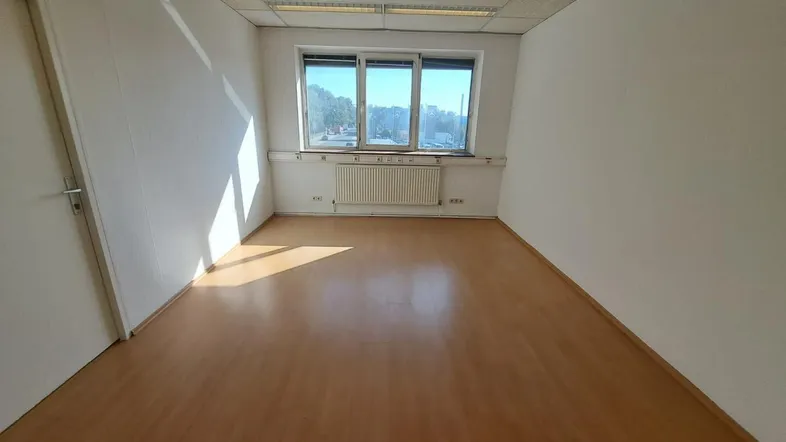 Heller, leerer Büroraum mit Laminatboden, weißen Wänden und großen Fenstern, die viel Tageslicht hereinlassen.