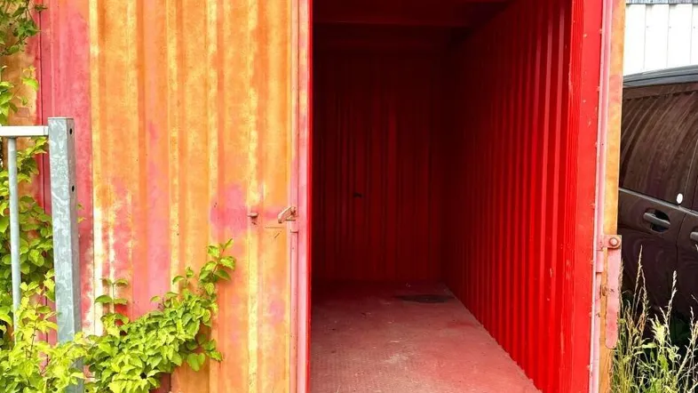 Rostiger roter Lagercontainer mit offener Tür, umgeben von Gras und Pflanzen auf einem unbefestigten Gelände.