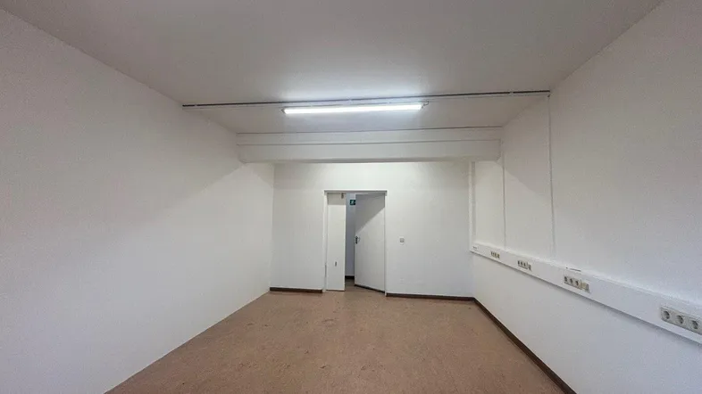 Geräumiger, leerer Büroraum mit hellen Wänden, Linoleumboden und Zugang zu einem weiteren Bereich.