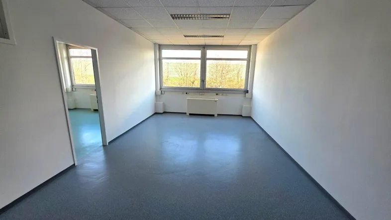 Langer, leerer Büroraum mit blauer Bodenfläche, weißen Wänden und großen Fenstern mit Weitblick.