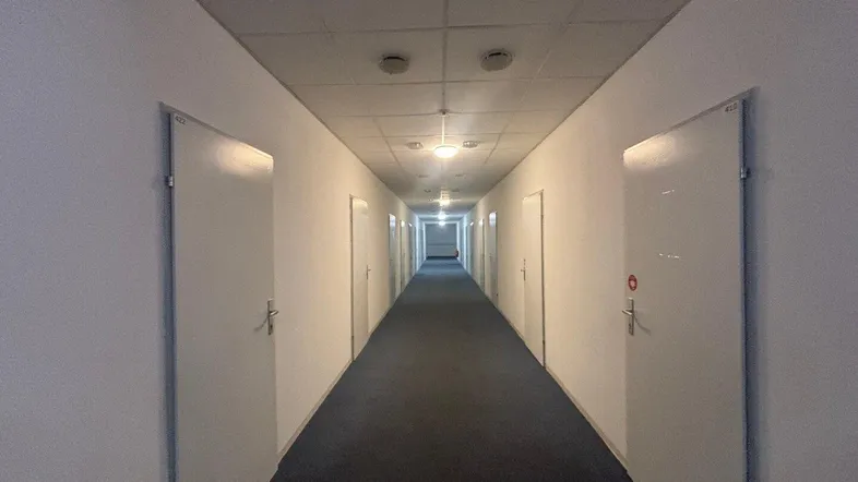 Langer, schmaler Flur mit Teppichboden und vielen Türen zu den einzelnen Wohneinheiten.