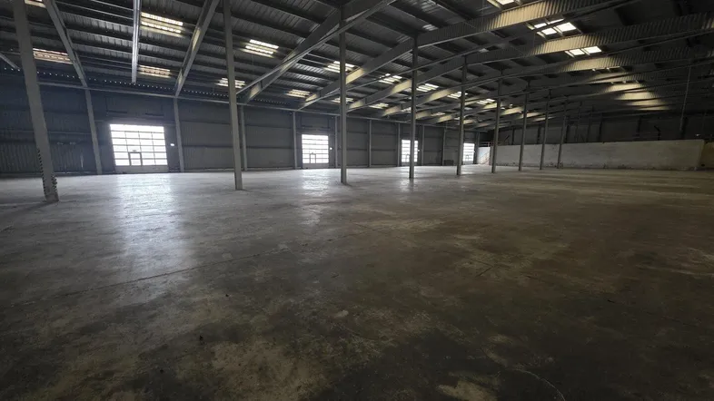 Große, leere Lagerhalle mit hoher Decke, Betonboden und Oberlichtern für natürliches Licht.