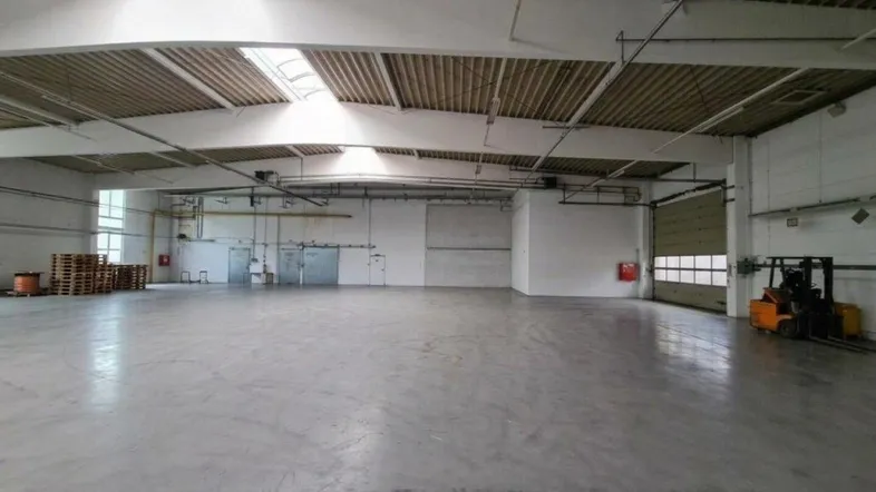 Große, leere Lagerhalle mit hohen Decken, Betonboden und viel Tageslicht durch Oberlichter und Fenster.