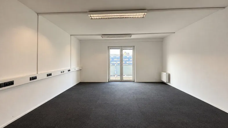 Geräumiger Büroraum mit dunklem Teppichboden, weißen Wänden und einer großen Glastür, die nach draußen führt.