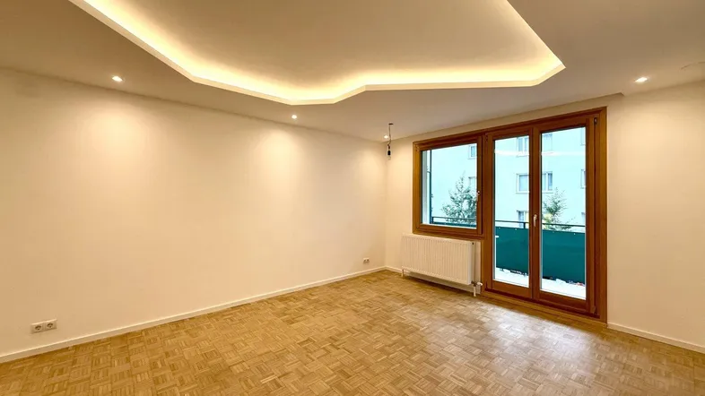 Helles Wohnzimmer mit Parkettboden, indirekter Deckenbeleuchtung und Zugang zum Balkon.