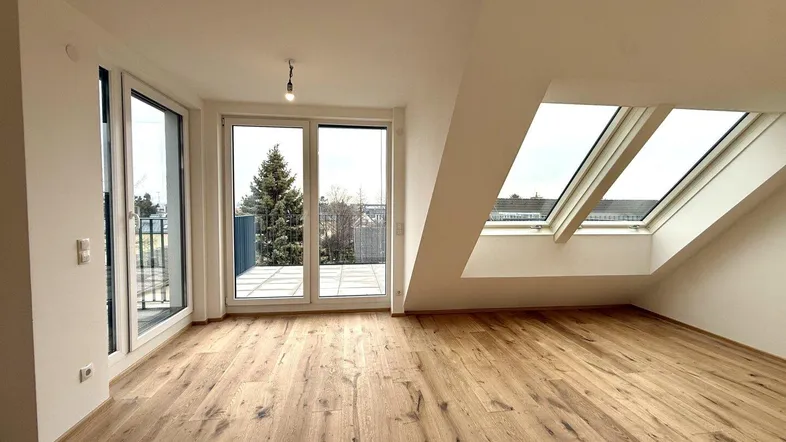Helles Zimmer mit Holzboden, großen Fenstern, Dachschräge und Zugang zu einer Terrasse.