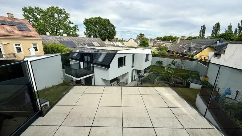 Große geflieste Terrasse mit Glasgeländer und Blick auf umliegende Häuser und Gärten.