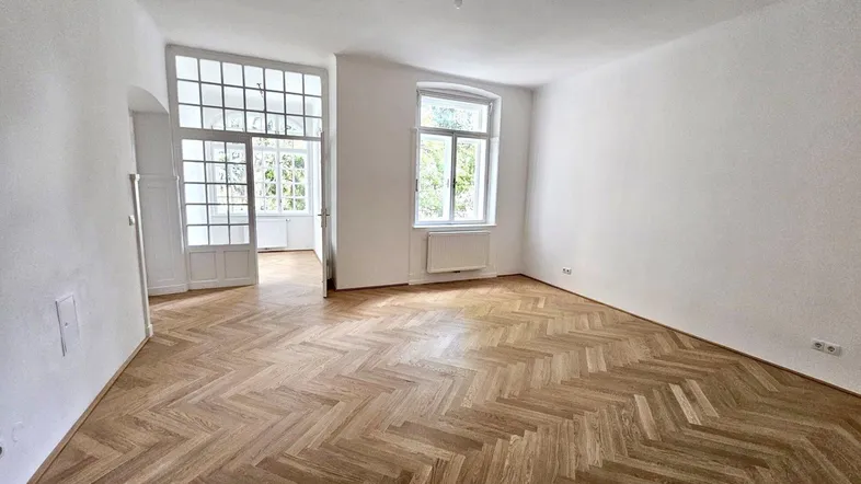 Helles Zimmer mit Fischgrätparkett und großen Fenstern, das viel Tageslicht hereinlässt.