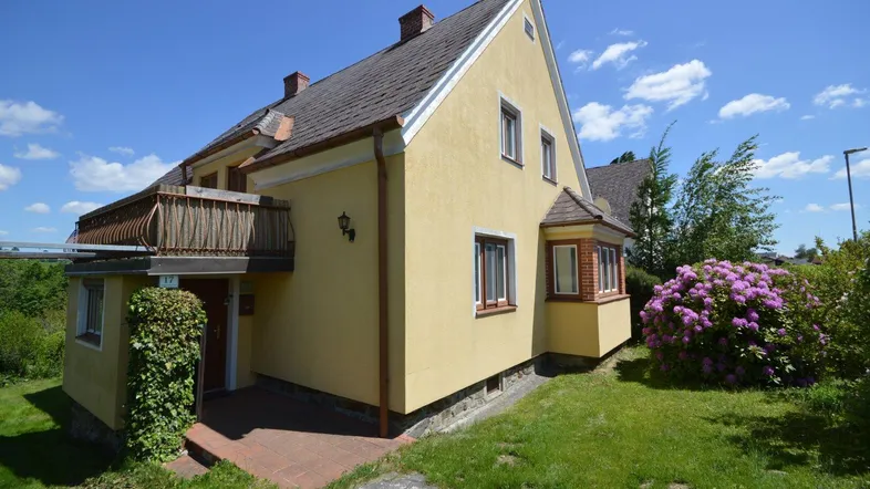 Gelbes Einfamilienhaus mit Balkon und blühendem Rhododendron im Vorgarten.