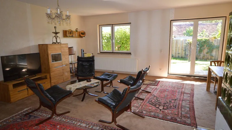 Gemütliches Wohnzimmer mit Teppichboden, traditionellem Kachelofen und Zugang zum Gartenbereich.