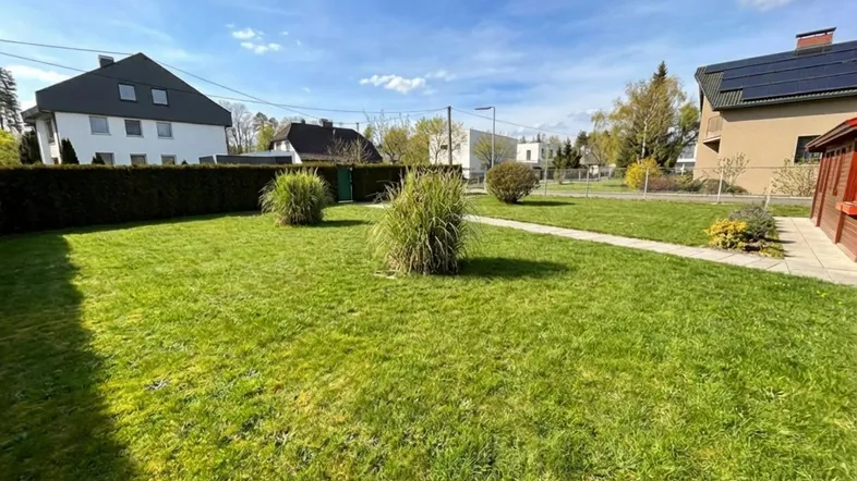 Gepflegter Gartenbereich mit Rasenfläche und Hecke, umgeben von Wohnhäusern unter blauem Himmel.
