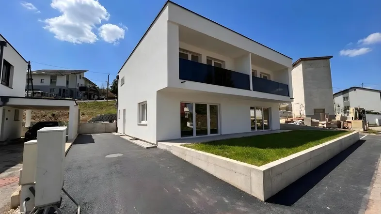Modernes Neubauhaus mit weißer Fassade, Balkonen und gepflegter Rasenfläche unter blauem Himmel.
