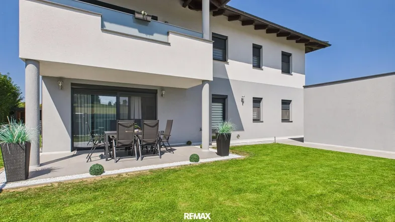 Moderne Hausfassade mit gepflasterter Terrasse, Gartenmöbeln und gepflegtem Rasenbereich.