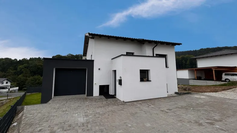 Modernes Einfamilienhaus mit weißer Fassade, schwarzer Garage und gepflasterter Auffahrt unter blauem Himmel.