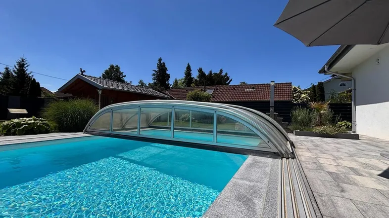 Großer Außenpool mit verschiebbarer Überdachung und gepflasterter Terrasse unter blauem Himmel.