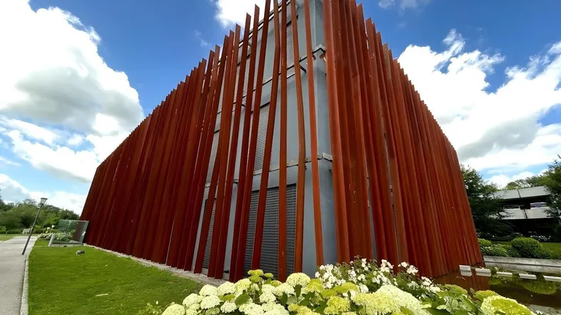 Modernes Gebäude mit rostfarbenen Lamellenfassaden und grüner Umgebung unter blauem Himmel.