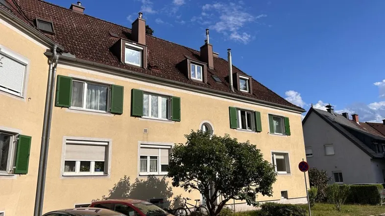 Gelbes Mehrfamilienhaus mit braunem Ziegeldach und grünen Fensterläden unter blauem Himmel.