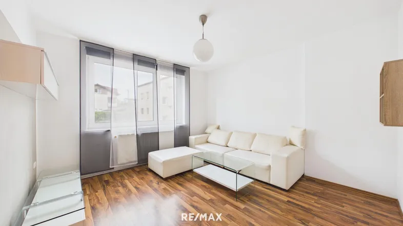 Helles Wohnzimmer mit Holzboden, weißem Sofa und großen Fenstern mit Vorhängen.