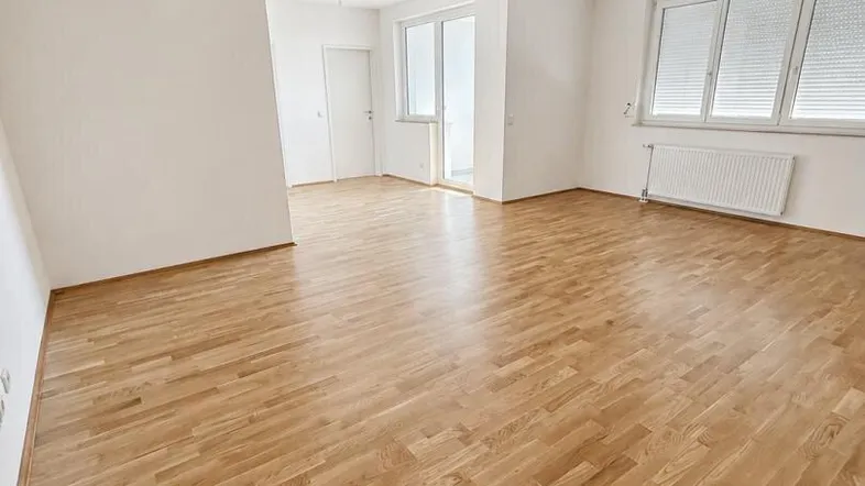 Helles, leeres Zimmer mit Parkettboden, weißen Wänden und Zugang zu einem Balkon oder einer Terrasse.