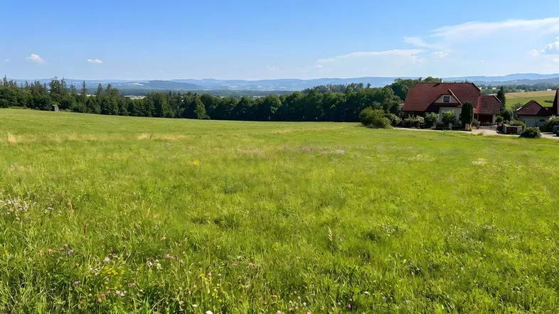 Weitläufiges grünes Feld mit Blick auf entfernte Häuser und eine hügelige Landschaft unter blauem Himmel.
