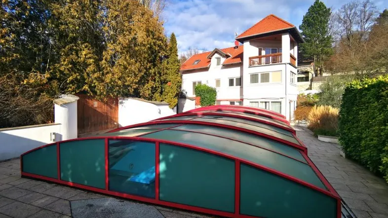 Außenansicht des Hauses mit einem überdachten Poolbereich und hohen Bäumen im Garten.