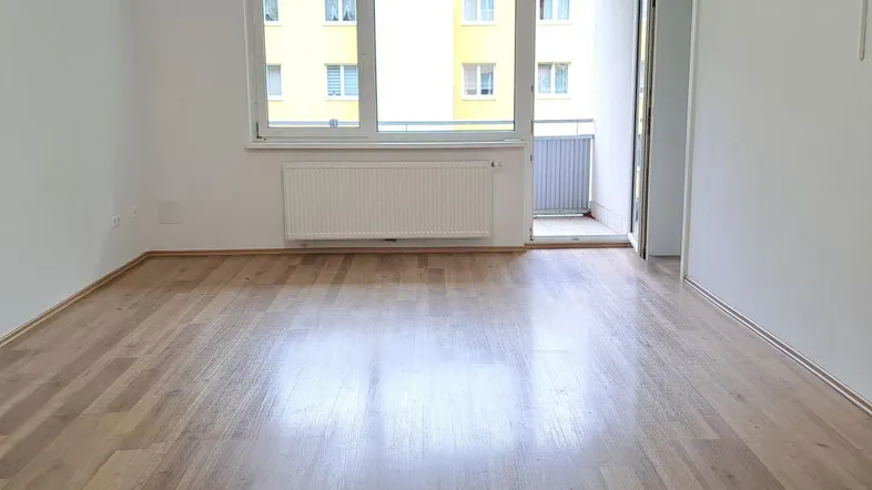 Helles Wohnzimmer mit Parkettboden, Heizkörper und Zugang zu einem kleinen Balkon.