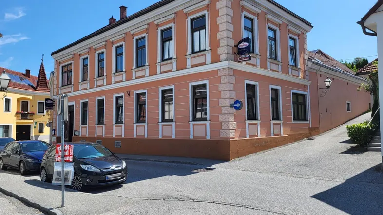 Historisches Gebäude mit pastellfarbener Fassade an einer Straßenecke, umgeben von parkenden Autos unter blauem Himmel.