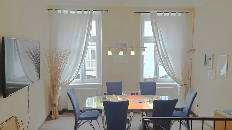 Heller Essbereich mit modernem Esstisch, blauen Stühlen und zwei großen Fenstern, die viel Tageslicht hereinlassen.