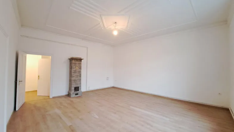 Geräumiges Zimmer mit hellem Laminatboden, Stuckdecke und traditionellem Kachelofen.