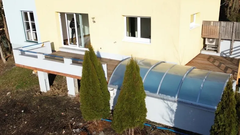 Modernes Haus mit gelber Fassade, großer Holzterrasse und überdachtem Poolbereich im Grünen.