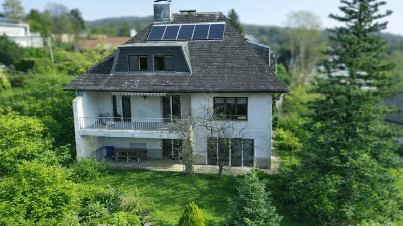 Freistehendes Haus mit Solaranlage auf dem Dach, umgeben von üppigem Grün und Bäumen.