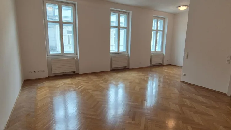 Helles Zimmer mit Fischgrätparkett, hohen Fenstern und klassischer Architektur.
