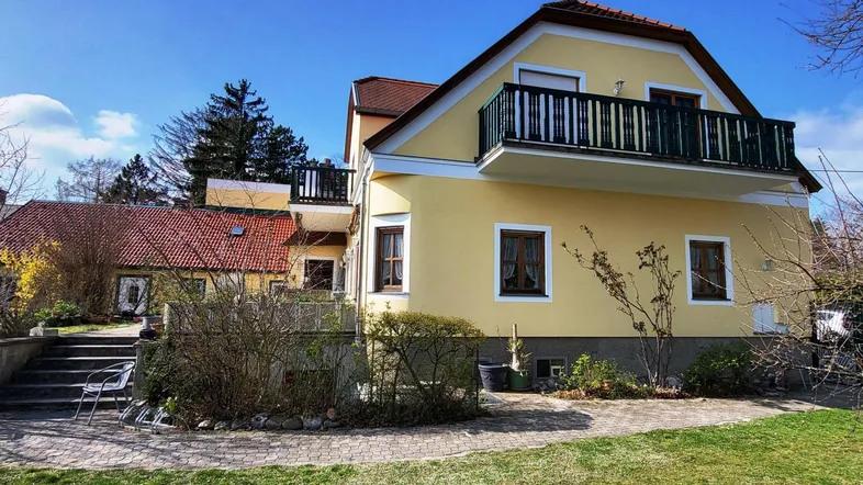 Gelbes Haus mit rotem Ziegeldach, Balkon und einem gepflegten Gartenbereich unter blauem Himmel.